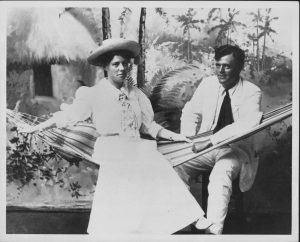 Black and white photo of a woman in a white early-20th-century dress with a hat and tie, sitting in a hammock, accompanied by a man in a white suit