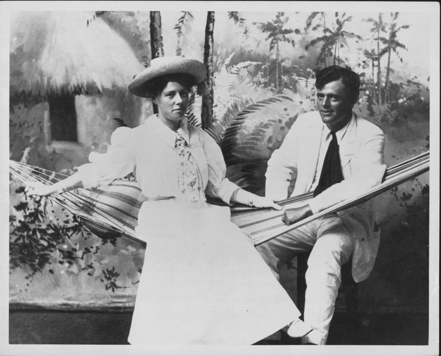 Black and white photo of a woman in a white early-20th-century dress with a hat and tie, sitting in a hammock, accompanied by a man in a white suit
