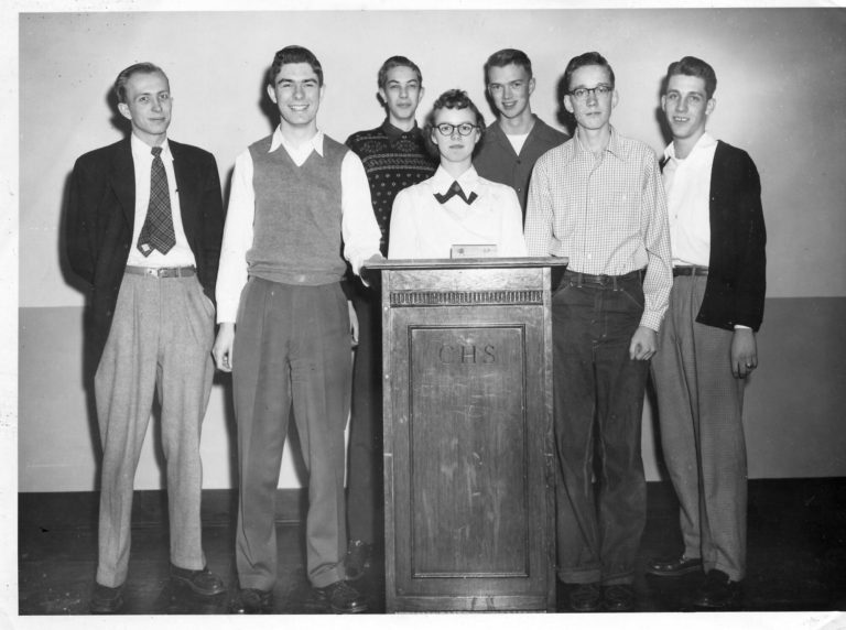 Black and white photo of a mid-century high school debate team. There are six skinny white men with various nerd accoutrements like glasses, sweater vests, and ill-fitting clothes, and one white woman with glasses standing behind a podium.