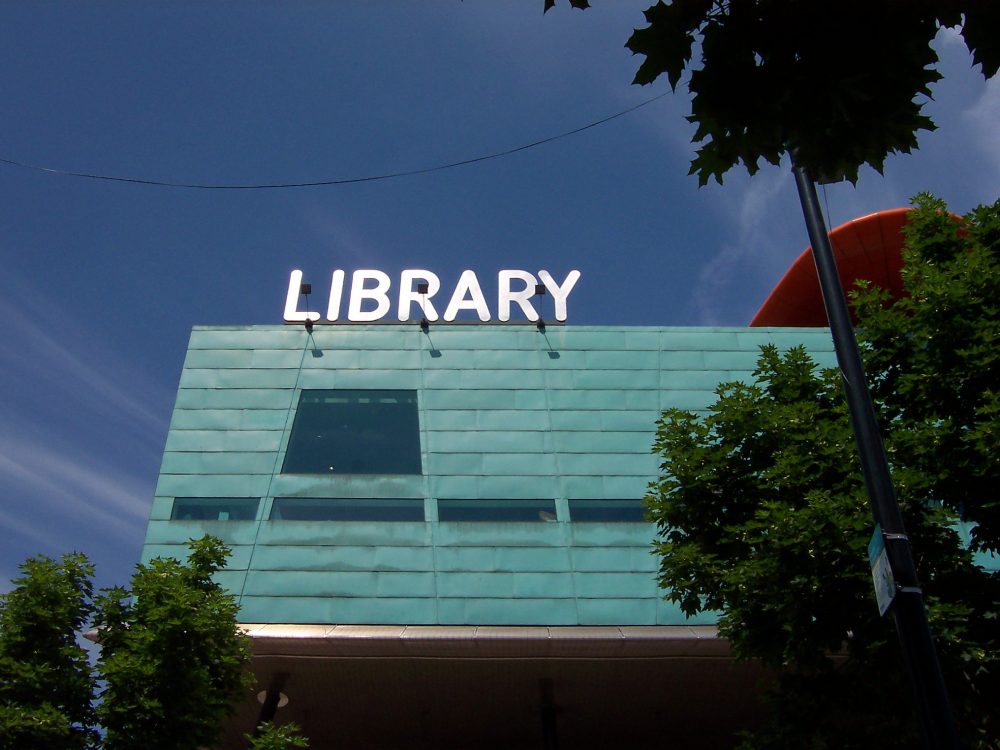 Boxy building with blue-green siding and an illuminated white sign reading LIBRARY on top
