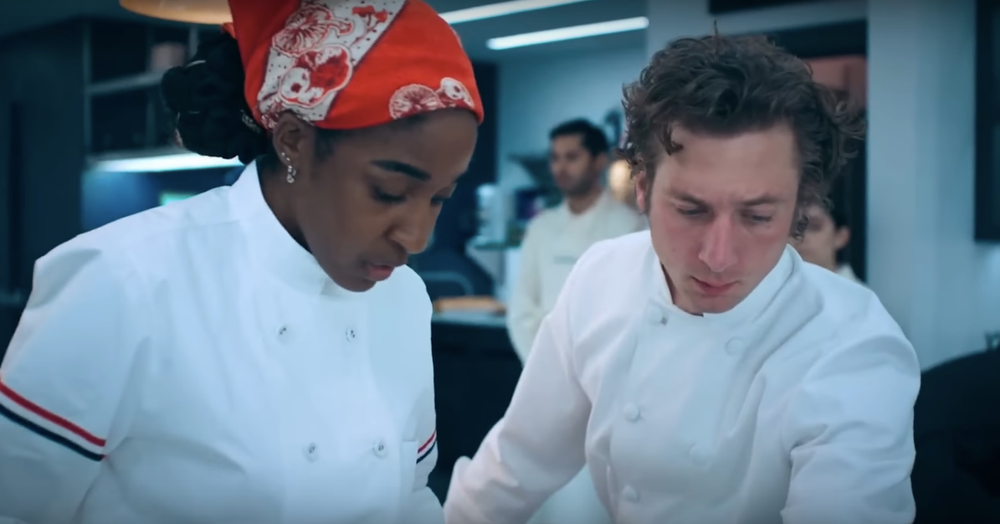 Carmy and Syd from "The Bear" chat in the kitchen wearing chef jackets