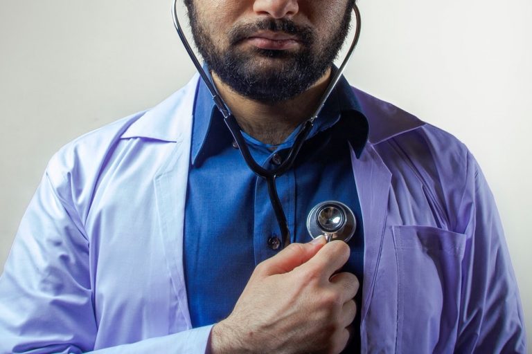 Man with brown skin and beard wearing lab coat and dress shirt, holding a stethoscope to his own chest
