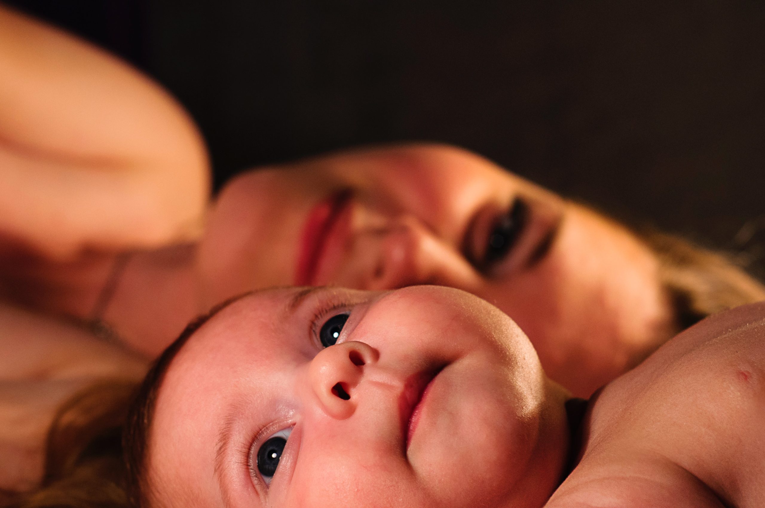A baby looks up while their mother smiles on from behind them.