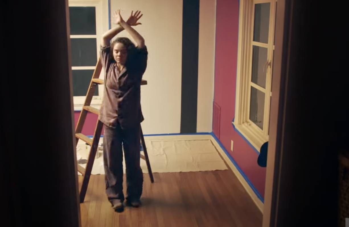 Standing in a nearly empty living room in front of a ladder, a standing woman stretches her arms