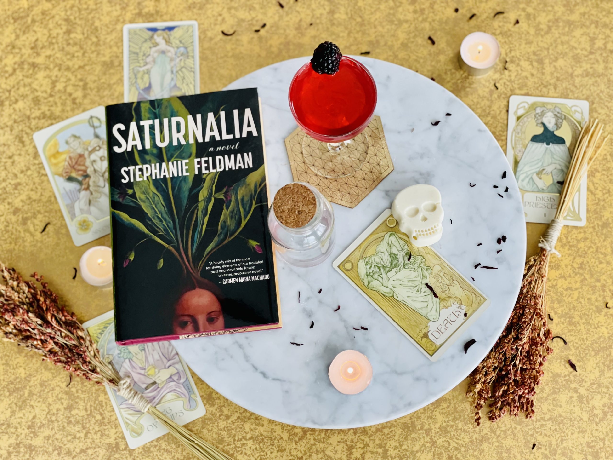 The book "Saturnalia" rests on a plate dotted with a candle, a tarot card, and a skull candy.