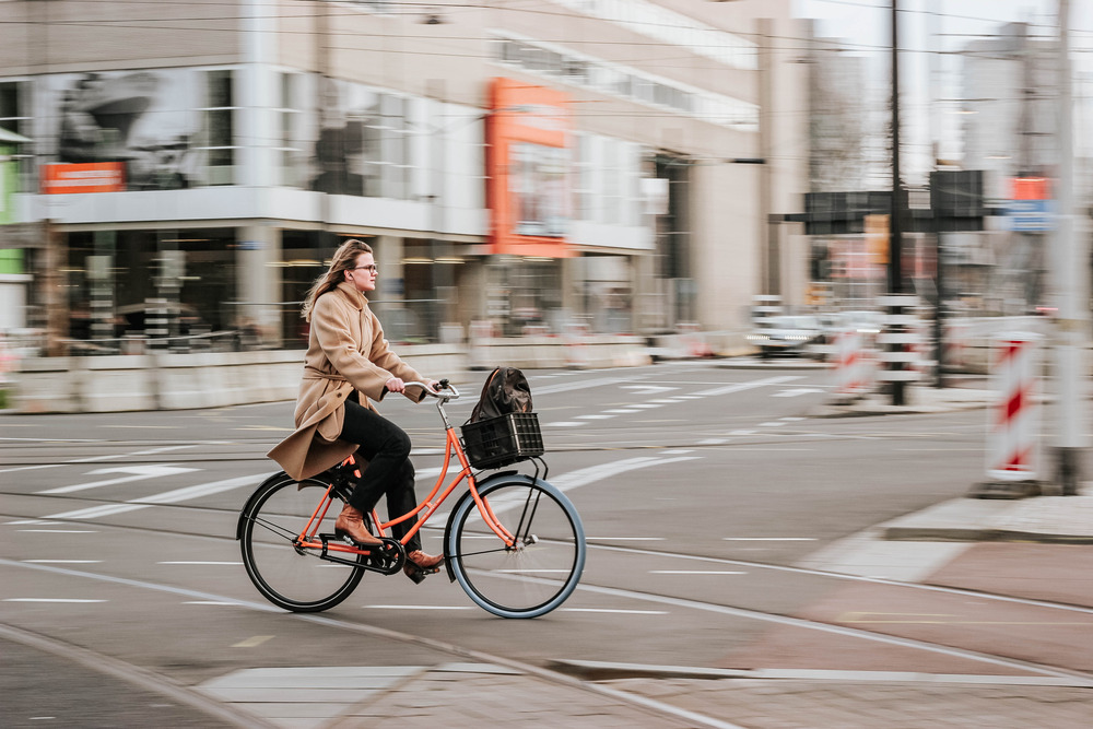 A woman in a beige coat rides an orange bike with a basket down an empty street.