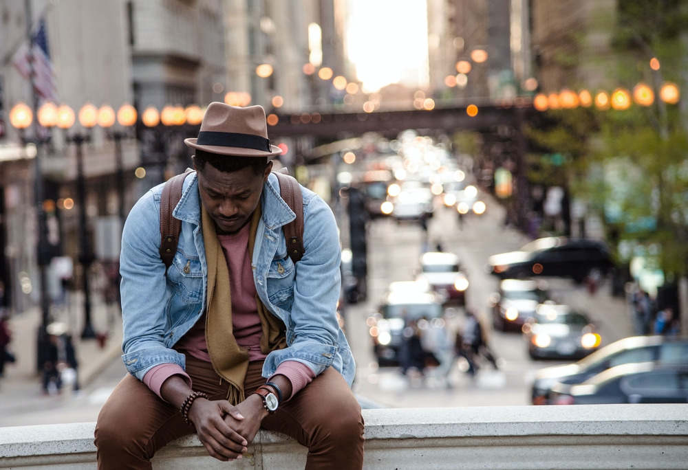 A man wearing a hat and denim shirt sits on a ledge while lights from the city glow behind him.