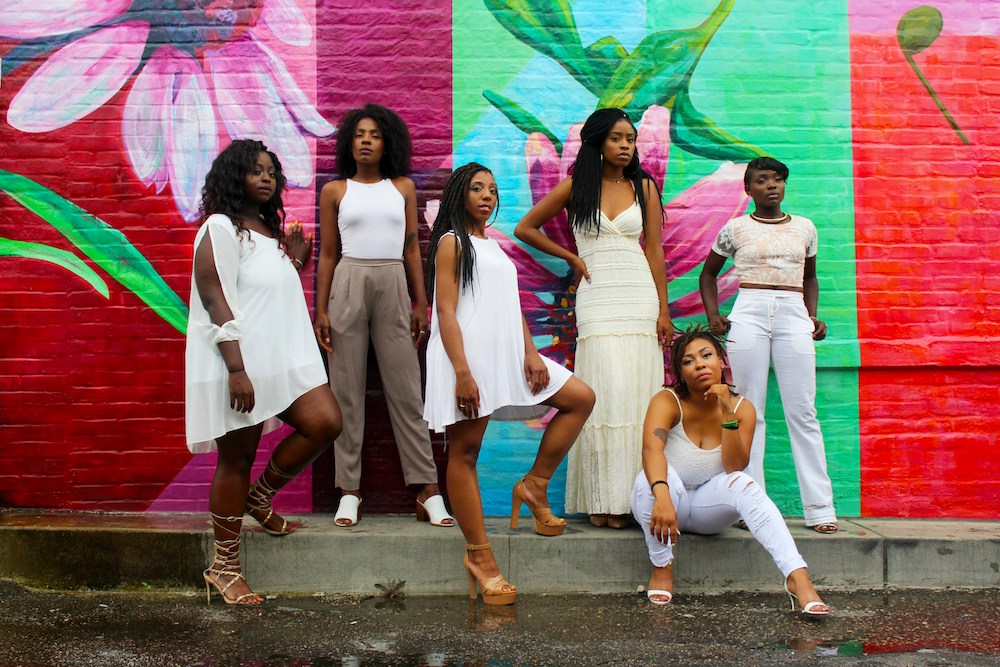 Six women wearing white and posing in front of a painted wall with floral graffiti