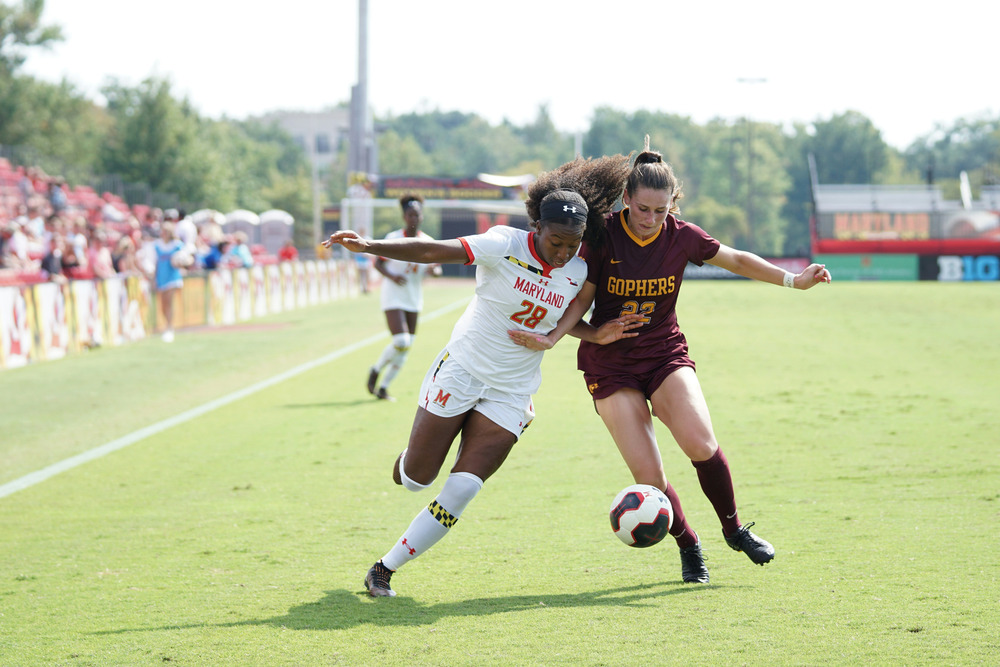 Two women--one in a white uniform, the other in a burgundy uniform--kick a soccer ball on a sports field
