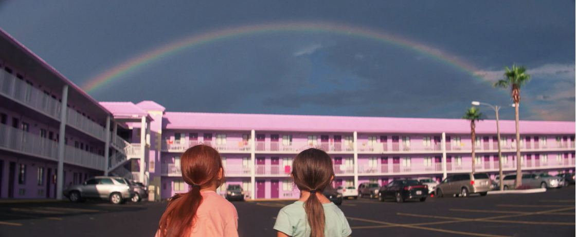 Two young girls stand in front of a pink hotel, looking up at a rainbow in the sky