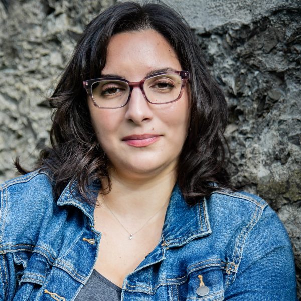 A queer author wearing a blue denim jacket with specs and a slight smile, staring into the camera