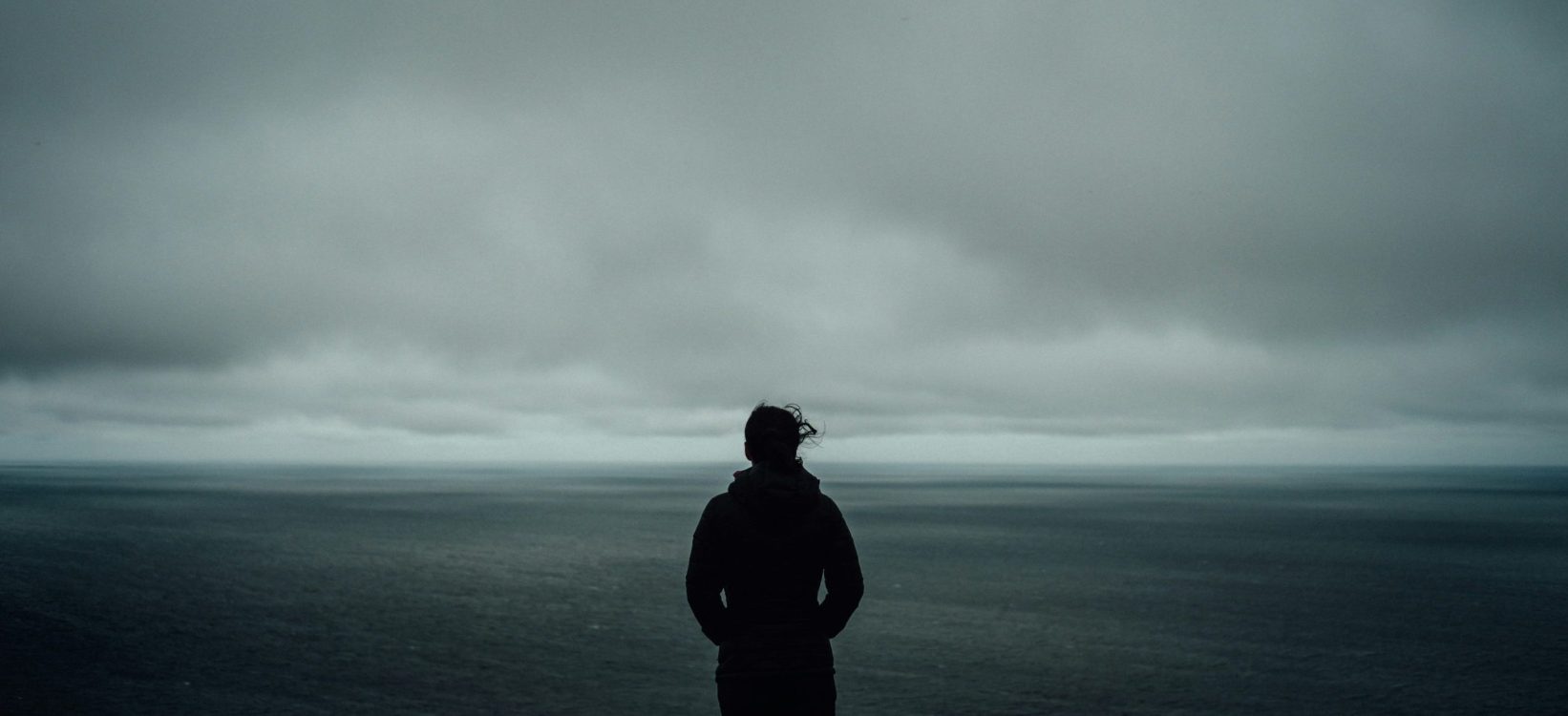 A person in a hoodie standing against a gloomy cloudy backdrop, their back to the viewer, hands in their pockets.