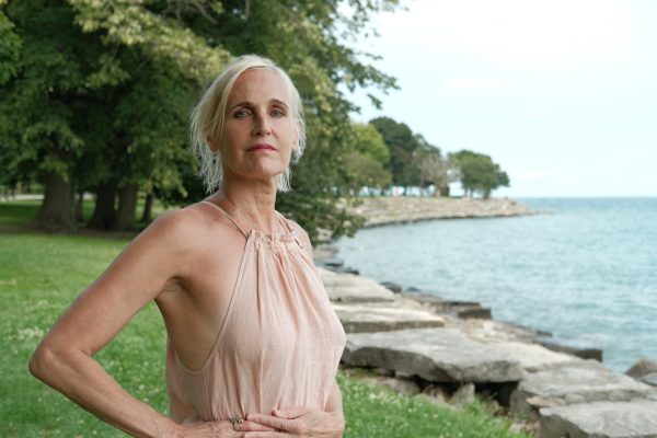 A person with white hair and beige dress against a background with trees and a water body looking into the camera